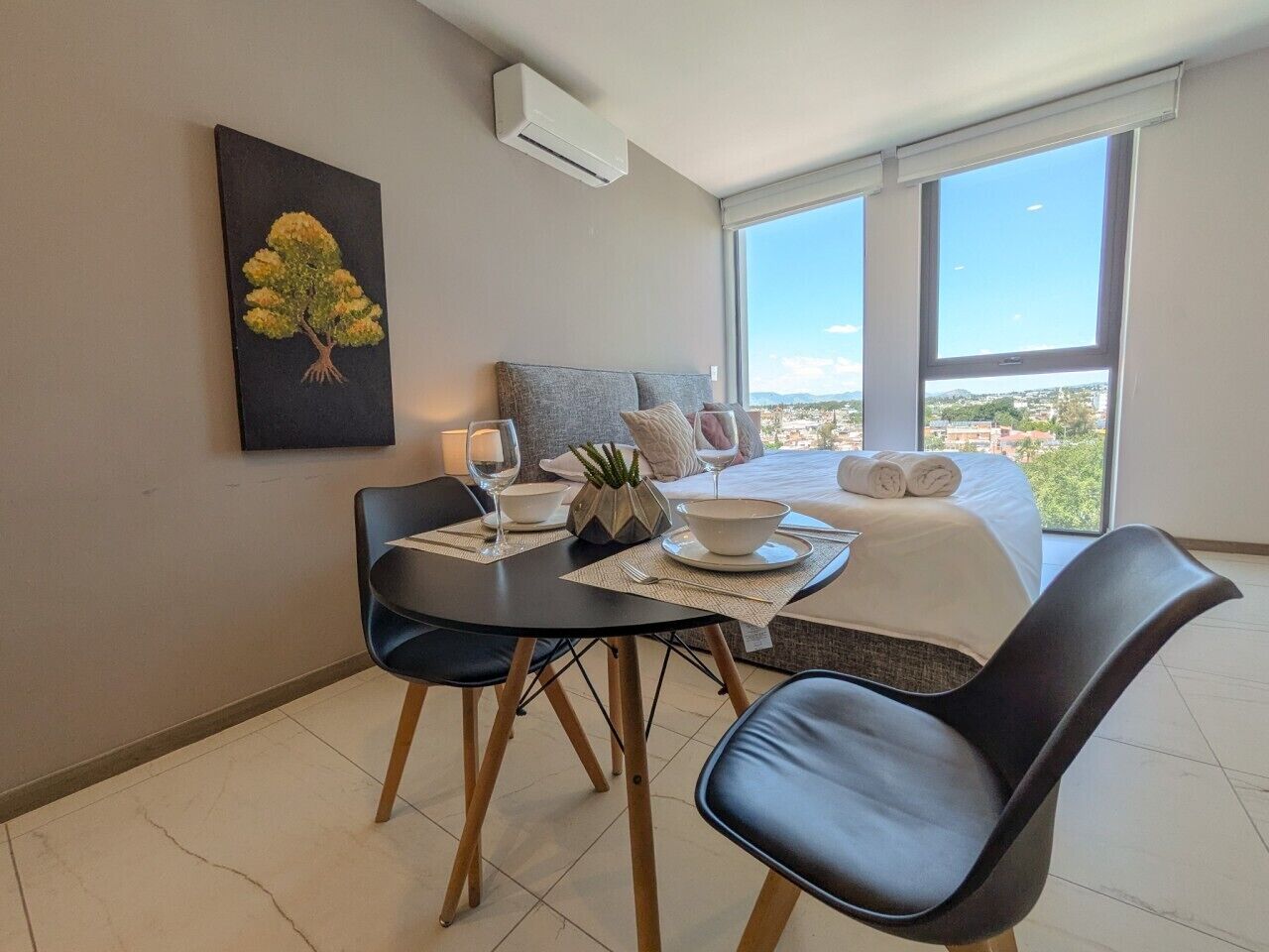 A modern hotel room with a dining table, chairs, a bed, and a large window offering a city view.