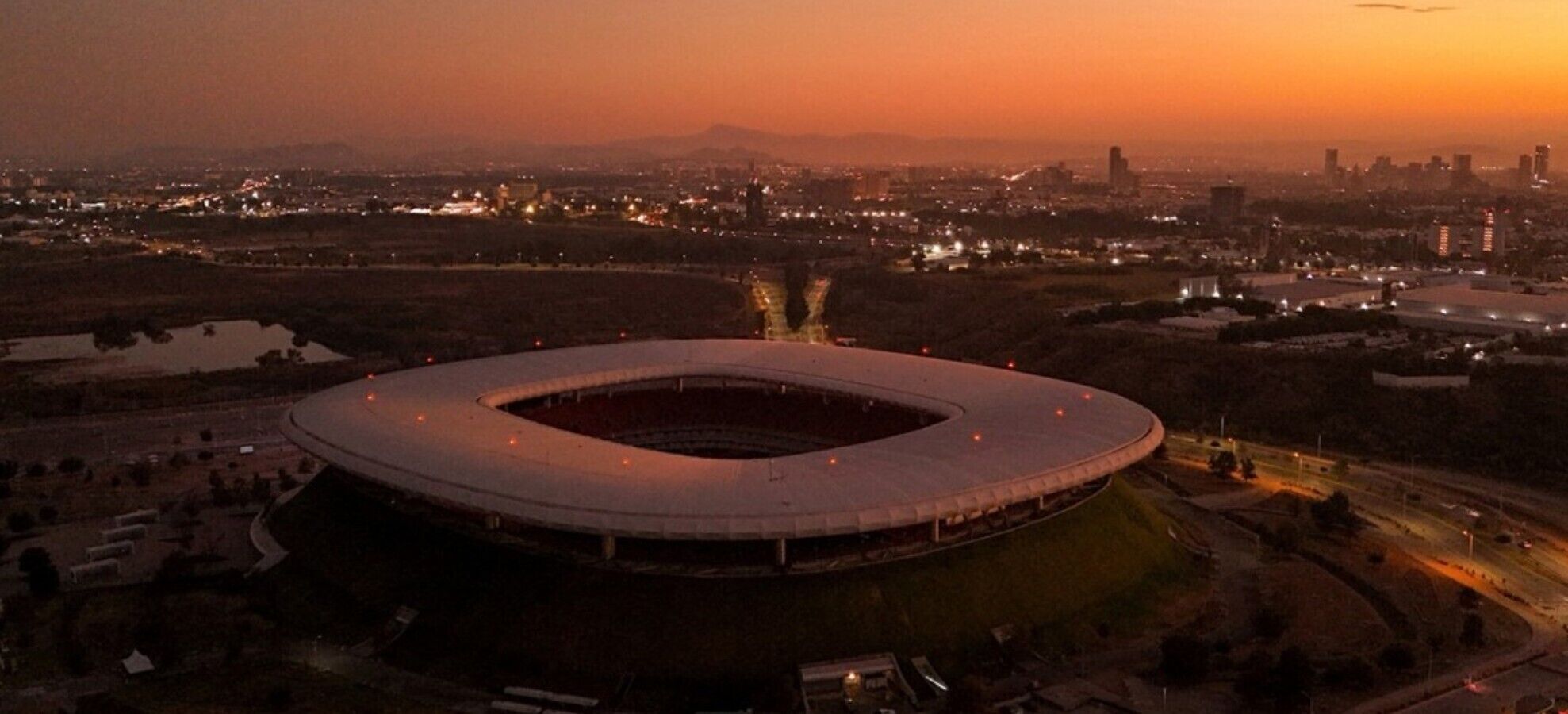 A stadium with a circular design, surrounded by a cityscape at dusk.