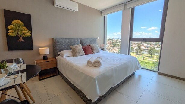 A modern bedroom with a large bed, a bedside table with a lamp, a small table with a glass of wine, and a view of the city through large windows. - Great New Apartment close to Andares A (Zapopan)