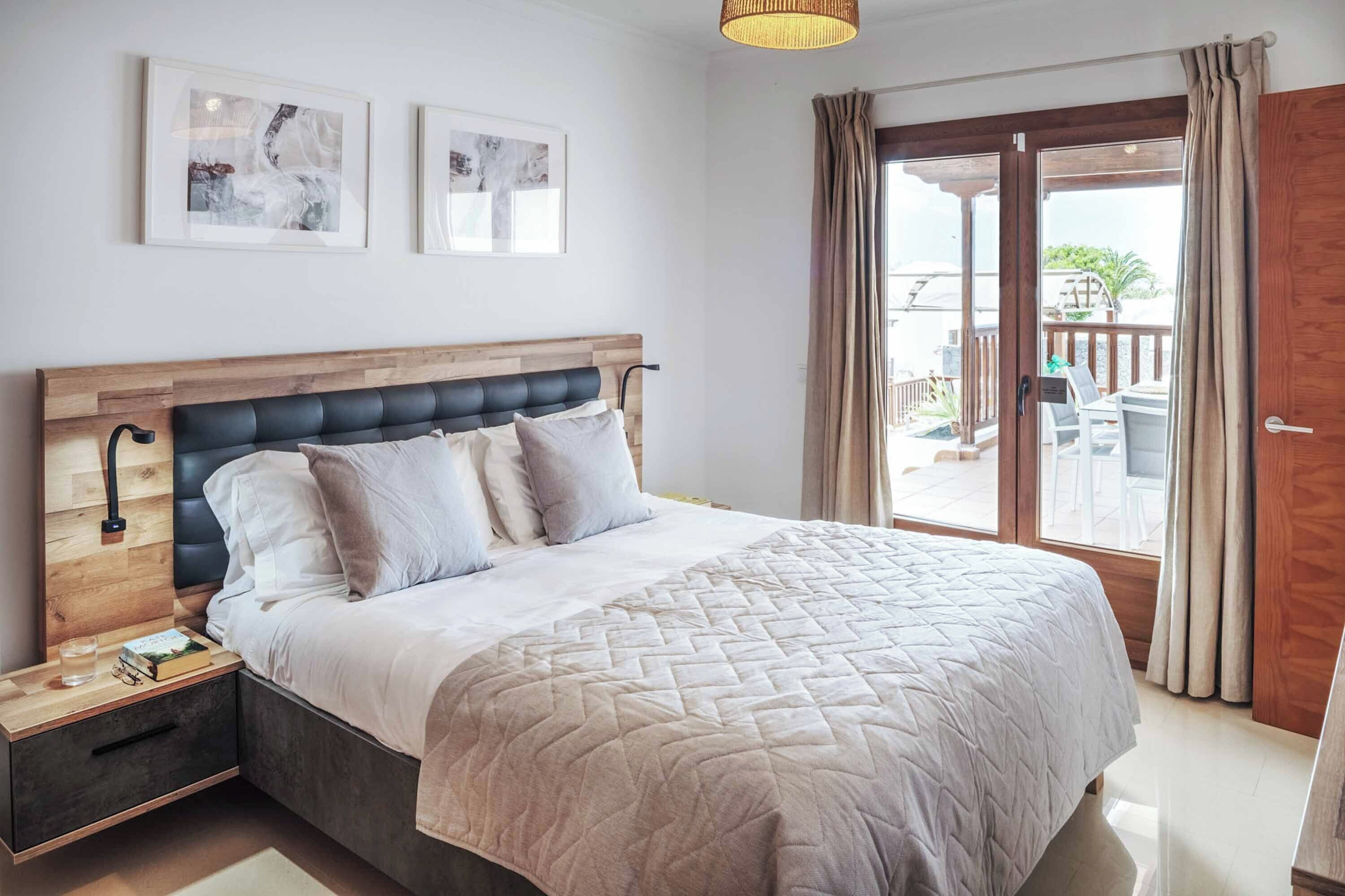 A bedroom with a wooden headboard, a bed with a quilted cover, a nightstand with a lamp, and a view of the outdoors through a sliding glass door.