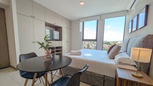 Ein modernes Hotelzimmer mit einem Bett, einem Esstisch mit Stühlen, einer kleinen Küchenzeile und einem großen Fenster mit Blick auf die Stadt.