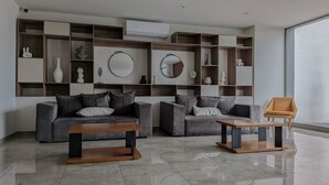 Uma sala de estar moderna com dois sofás, uma mesa de centro e uma mesa lateral. Há prateleiras com objetos decorativos e um ar condicionado na parede.