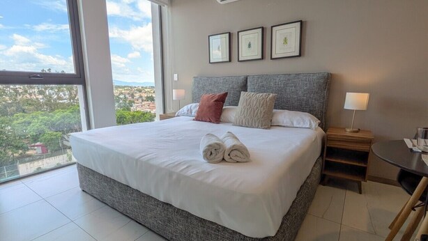 Ein Hotelzimmer mit einem großen Bett, einem Nachttisch, einer Lampe und Blick auf die Stadt.
