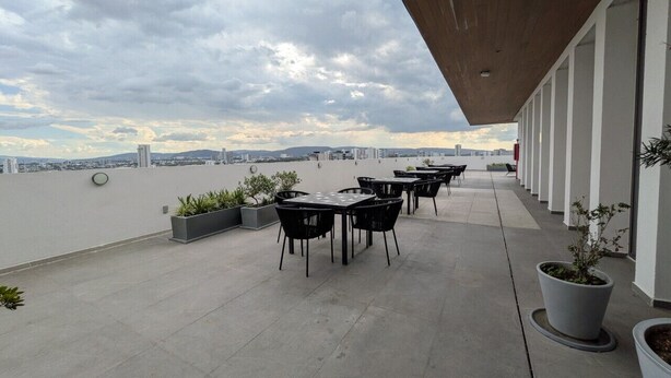 Terraço ao ar livre com mesas e cadeiras, plantas em vasos e vista para a cidade.