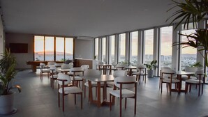 A spacious dining area with multiple tables and chairs, large windows offering a city view, and potted plants. - New Loft near Andares B (Zapopan)