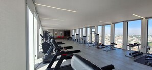 Un gimnasio con diverso equipo de ejercicio, amplias ventanas y vistas a la ciudad.