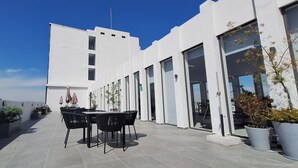 Une terrasse sur le toit, avec une table et des chaises, des plantes en pot et un bâtiment moderne doté de grandes fenêtres et d’un toit plat.