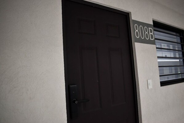 Une porte sombre, avec le numéro « 808B » au-dessus, une fenêtre avec des stores horizontaux et un interrupteur sur le mur.