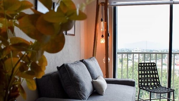 Ein modernes Wohnzimmer mit einer Couch, einem Esstisch und einem Sessel. Eine Pflanze in einer Vase, eine Pendelleuchte und Blick auf die Stadt durch große Fenster.