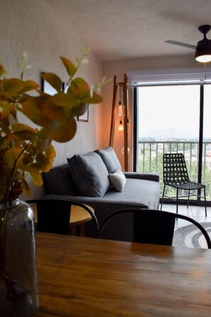 Un salon moderne avec un canapé, une table à manger et une chaise. On y trouve une plante dans un vase, un luminaire suspendu et une vue sur la ville à travers de grandes fenêtres.