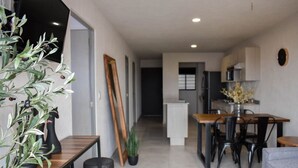 Eine moderne Küche mit weißen Schränken, eine Essecke mit einem Holztisch und schwarzen Stühlen sowie ein Wohnbereich mit einem grauen Sofa und einem Holztisch.