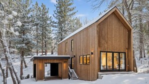 A wooden cabin with a large glass door and windows, surrounded by snow-covered trees.
