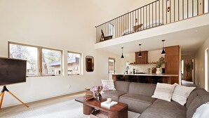 A spacious living room with a large sectional sofa, a wooden coffee table, and a flat-screen TV on a stand. A balcony with a railing is visible above the sofa.