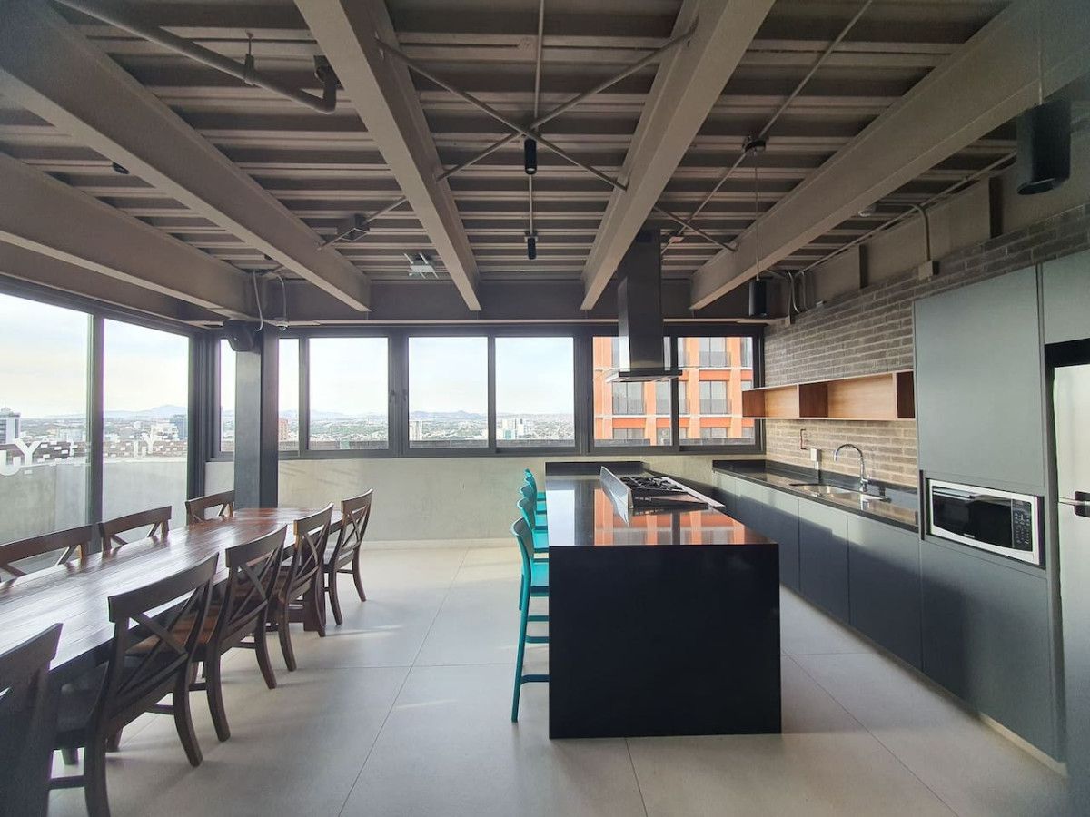 A modern kitchen with a large island, a dining area with wooden tables and chairs, and large windows offering a city view.