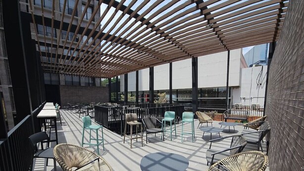 Un espace extérieur aménagé avec des chaises et des tables en osier, une pergola en bois et une vue sur un bâtiment moderne en arrière-plan.