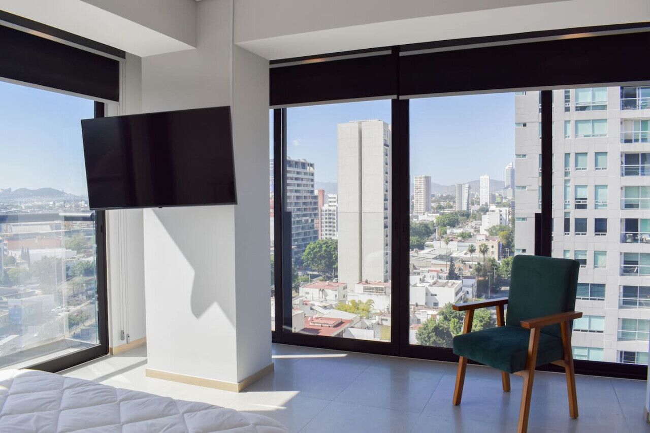 A modern hotel room with a large window, a flat-screen TV, and a green chair.