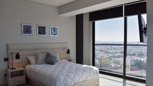 Un dormitorio moderno con una cama grande, mesitas de noche y vistas a la ciudad a través de amplios ventanales.
