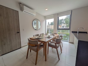 Un comedor moderno con una mesa de madera dispuesta para cuatro personas, sillas alrededor, un espejo grande y vistas al exterior a través de una puerta corredera de vidrio.