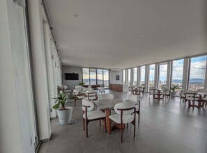 Un comedor espacioso con varias mesas y sillas, ventanas amplias que ofrecen vistas de la ciudad y un televisor de pantalla plana montado en la pared.