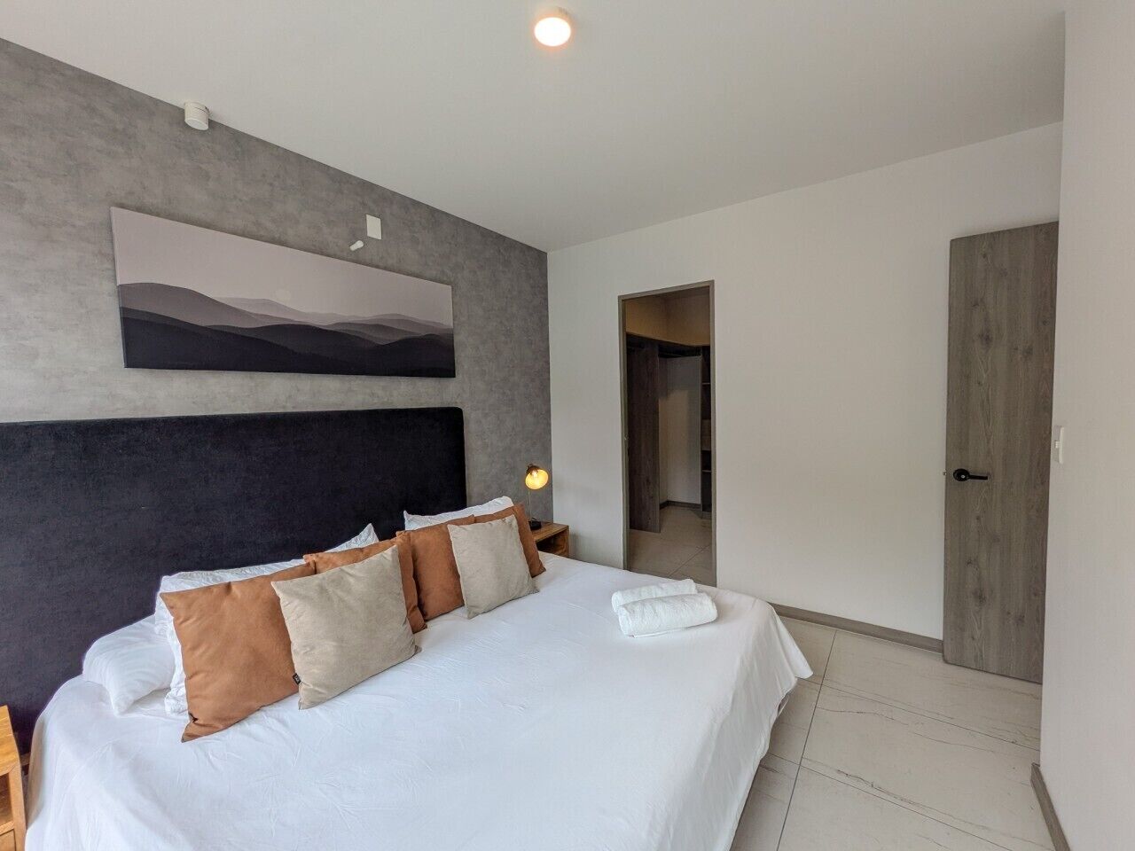 A modern hotel room with a large bed, a headboard, a painting on the wall, a bedside lamp, and a wooden door.
