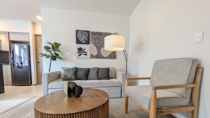 Een moderne woonkamer met een bank, een houten salontafel en een fauteuil. Er staat een lamp op een bijzettafeltje en er hangt kunst aan de muur.