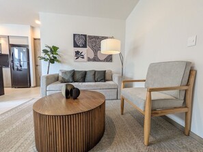 Una sala moderna con sofá, una mesa de centro de madera y una silla. Hay una lámpara en una mesita auxiliar y una obra de arte en la pared.