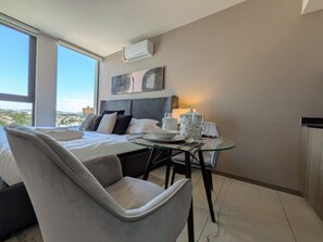 A hotel room with a bed, a dining table with chairs, and a large window offering a city view.