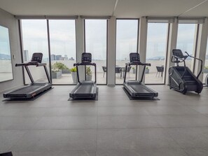 Un gimnasio con cintas de correr y una escalera mecánica, ventanas amplias y vistas a un paisaje urbano.