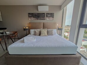 A hotel room with a large bed, a desk, chairs, and a view of the city through large windows. - Amazing new apartment near Andares 1209B (Zapopan)