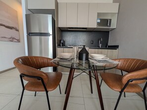 Cocina moderna con refrigerador de acero inoxidable, microondas y cafetera sobre la encimera. En el centro, una mesa de vidrio con dos sillas.