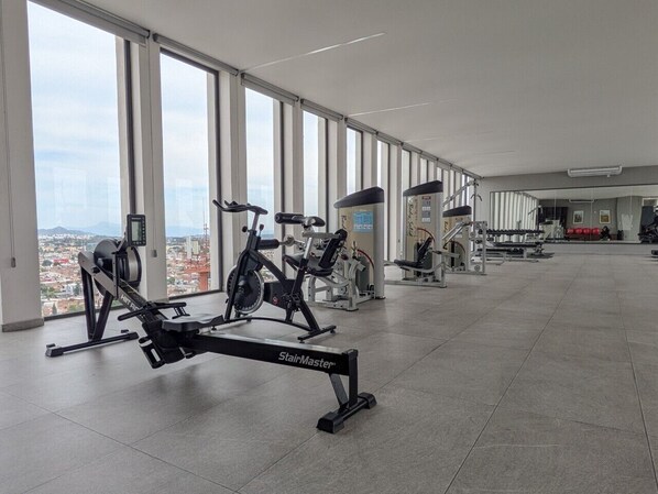 Un gimnasio con diverso equipo de ejercicio, amplias ventanas y vista a un paisaje urbano.