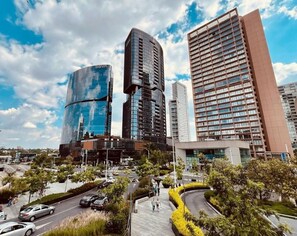 A modern urban area with high-rise buildings, a curved glass structure, and a pedestrian walkway with yellow landscaping.