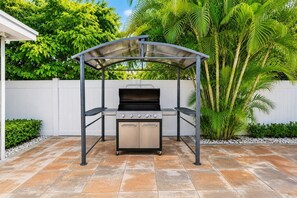 An outdoor grill with a metal canopy, surrounded by a patio with stone tiles and a white fence.