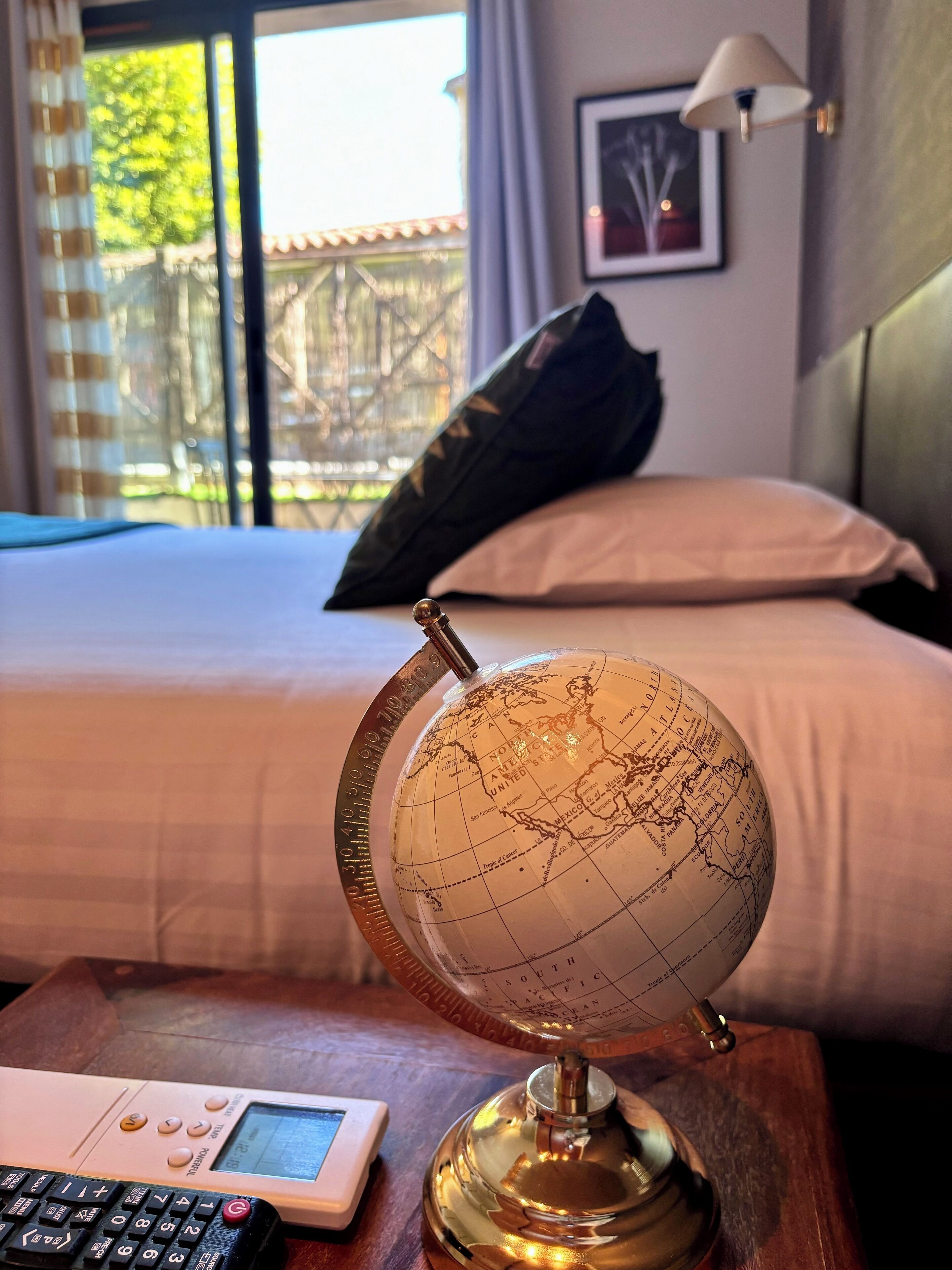 Ein Globus auf einem Holztisch in einem Hotelzimmer, im Hintergrund ein Bett, Kissen und eine Lampe.