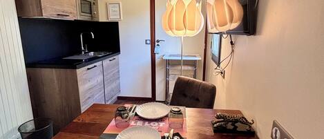 A dining area with a wooden table set for two, a television mounted on the wall, and a kitchen area with a microwave and a refrigerator.