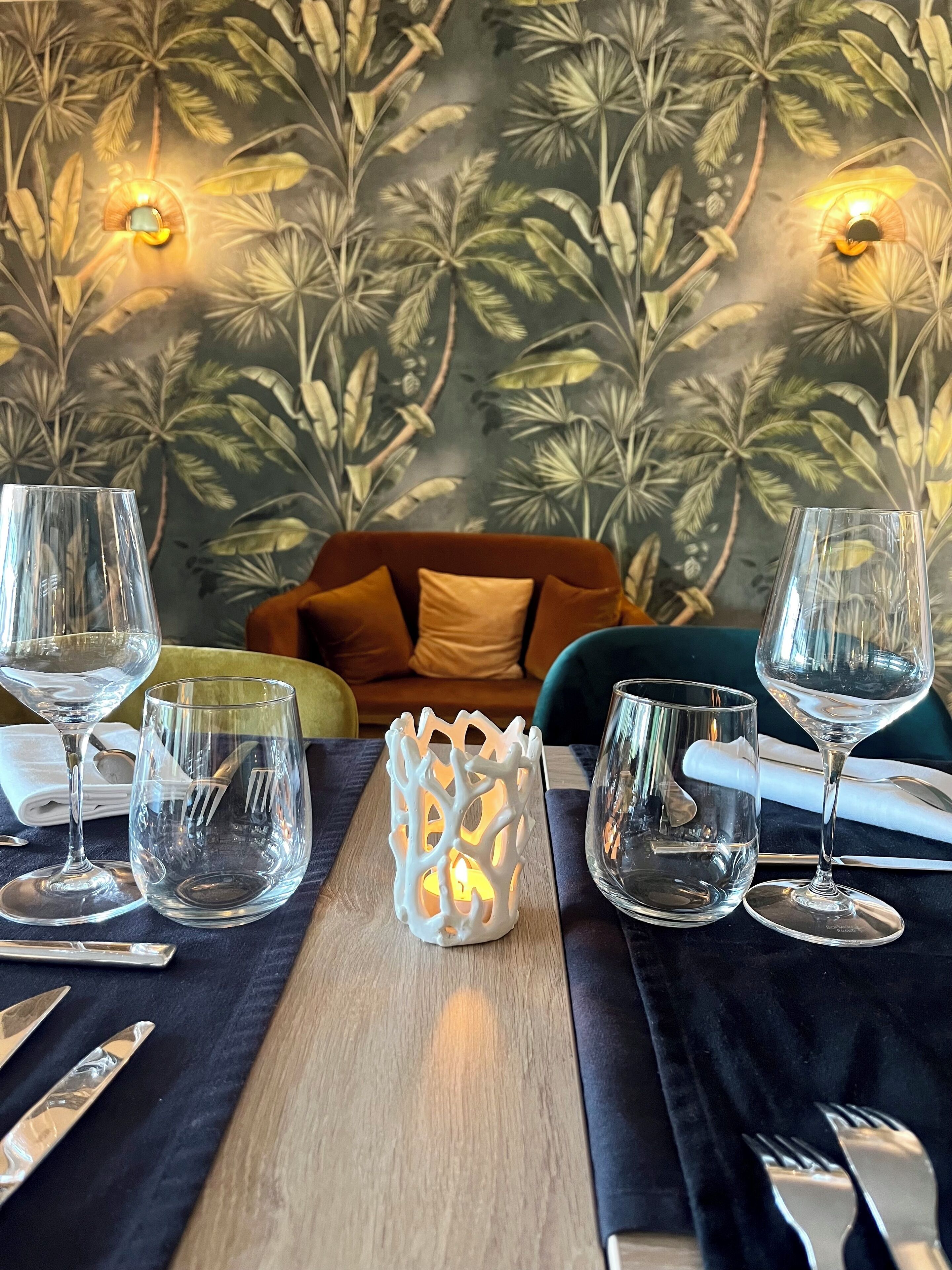 A dining table set with clear glassware, a white candle holder, and a tropical leaf-patterned wall in the background.