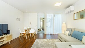 A modern living room with a flat-screen TV, a sofa, a dining table with chairs, and a large window with sheer curtains. - Bombarda Patio Oasis by LovelyStay (Porto)