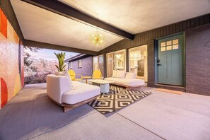 An outdoor patio area with a white sofa, yellow chairs, and a coffee table. There is a geometric rug on the floor, and a green door is visible in the background. - For Families and Large Groups! Hot Tub, Turf Yard, Outdoor Dining and Games (Wheat Ridge)