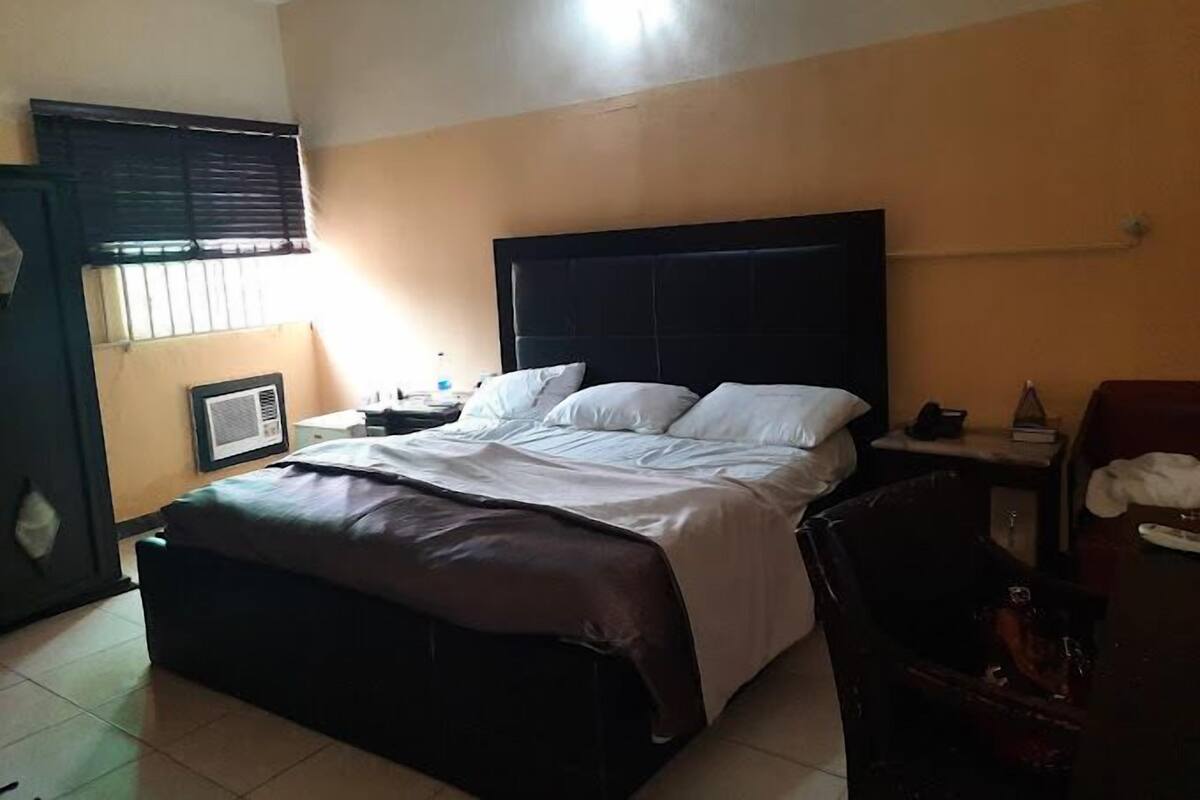 A hotel room with a large bed, a bedside table, a chair, and a window with blinds.