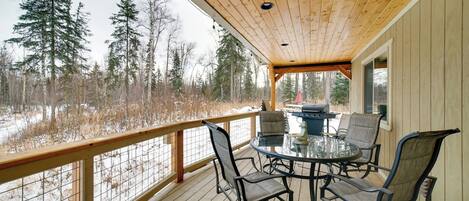 A wooden deck with a glass table and chairs, overlooking a snowy landscape with trees.