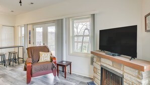 A living room with a stone fireplace, a large flat-screen TV, a comfortable armchair with a pillow, and a dining area with a bar stool. - Apt w/ Lake Views - Day Trip to Virginia Tech! (Radford)