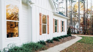 A white house with wooden shutters, a pathway, and a wooded area in the background.