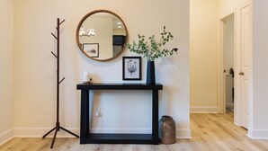 A hallway with a black console table, a mirror, a framed picture, a vase with green plants, a coat rack, and a door leading to another room.