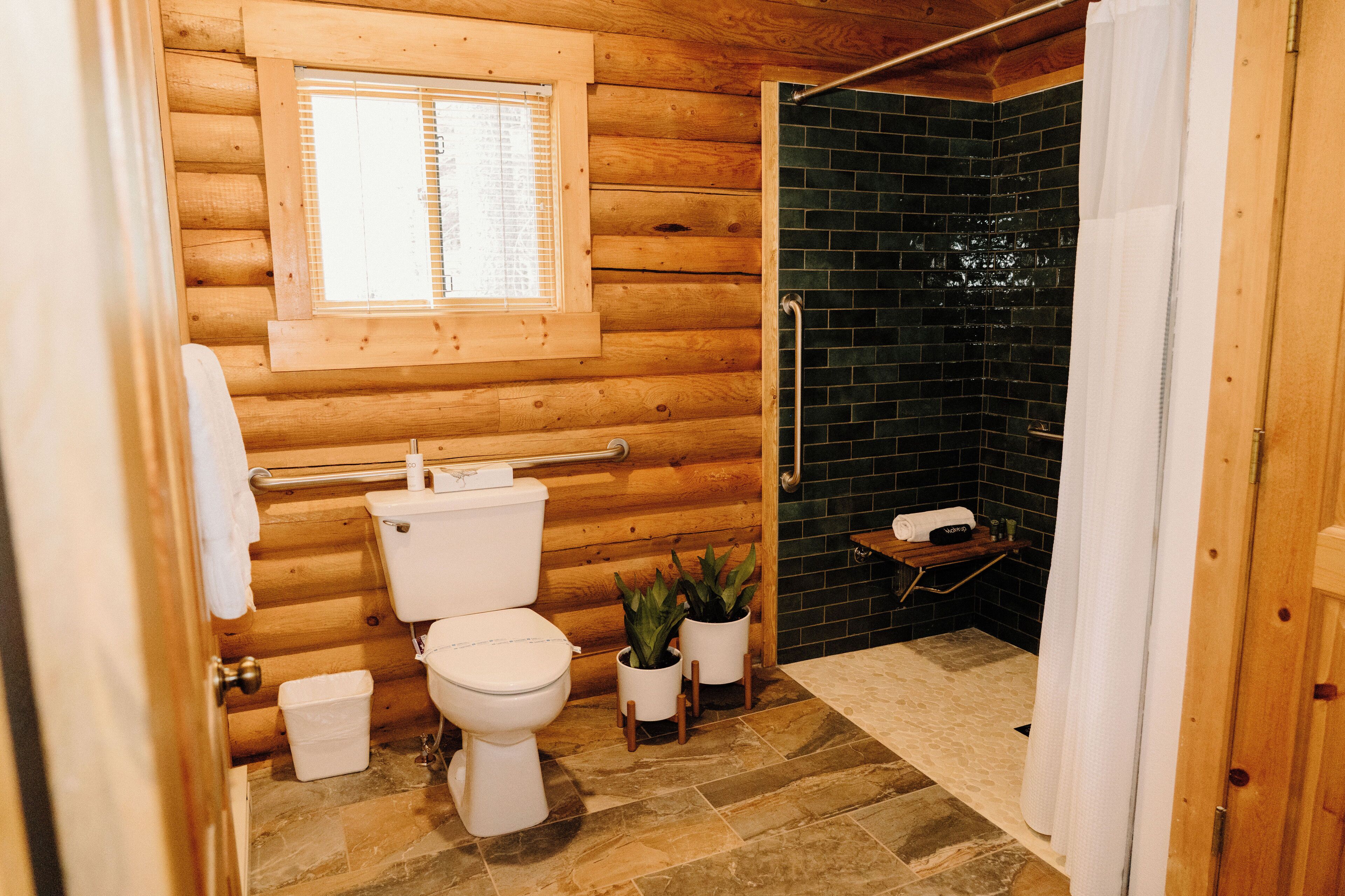 A wooden cabin bathroom with a toilet, two potted plants, a window, and a shower area with a tiled wall.