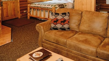 A log cabin interior with a wooden ceiling, a bed with a patterned blanket, a brown leather couch, and a small table with a book and remote control.