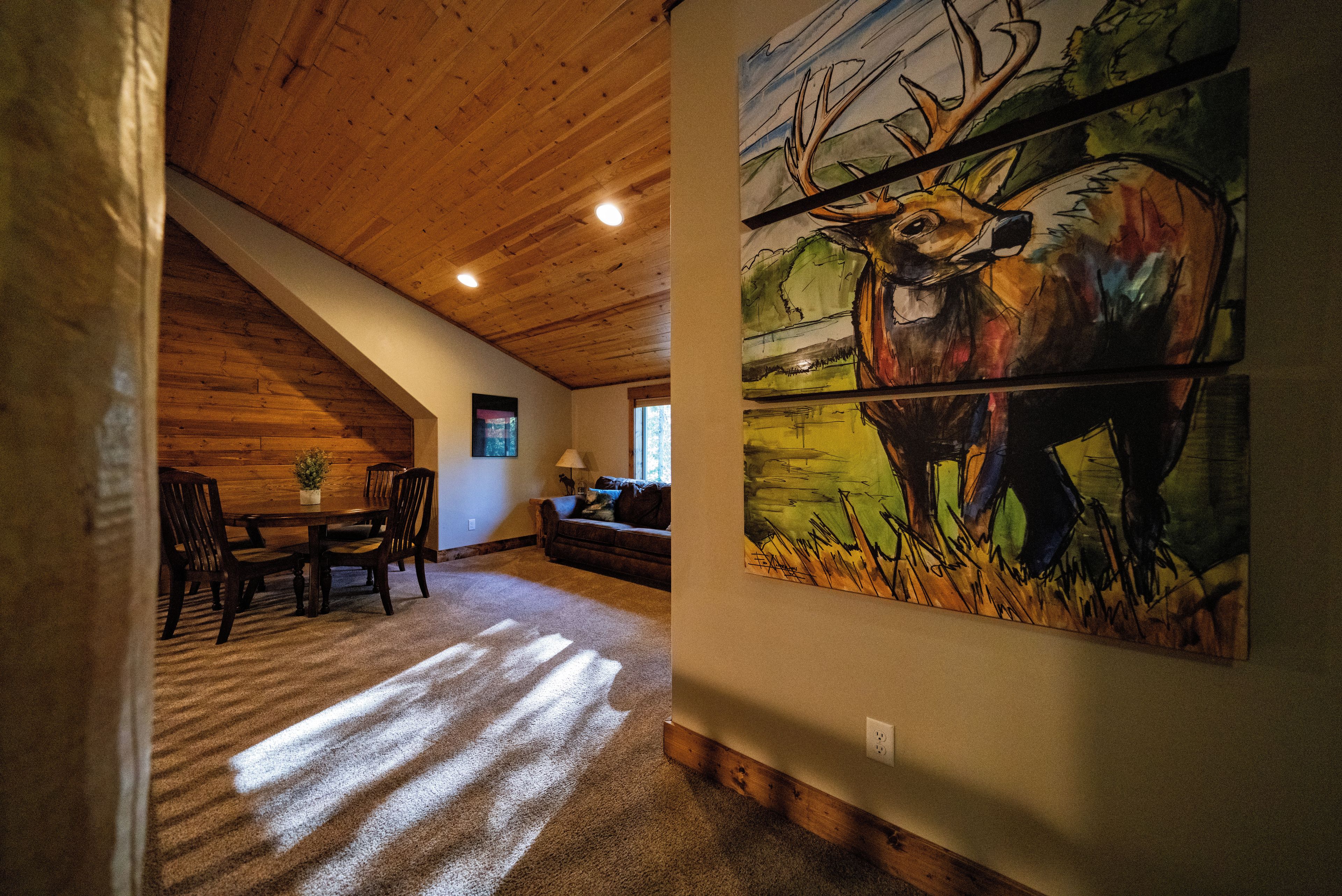 Een kamer met een schuine kap, een eethoek met een tafel en stoelen, en een schilderij van een hert op de muur.