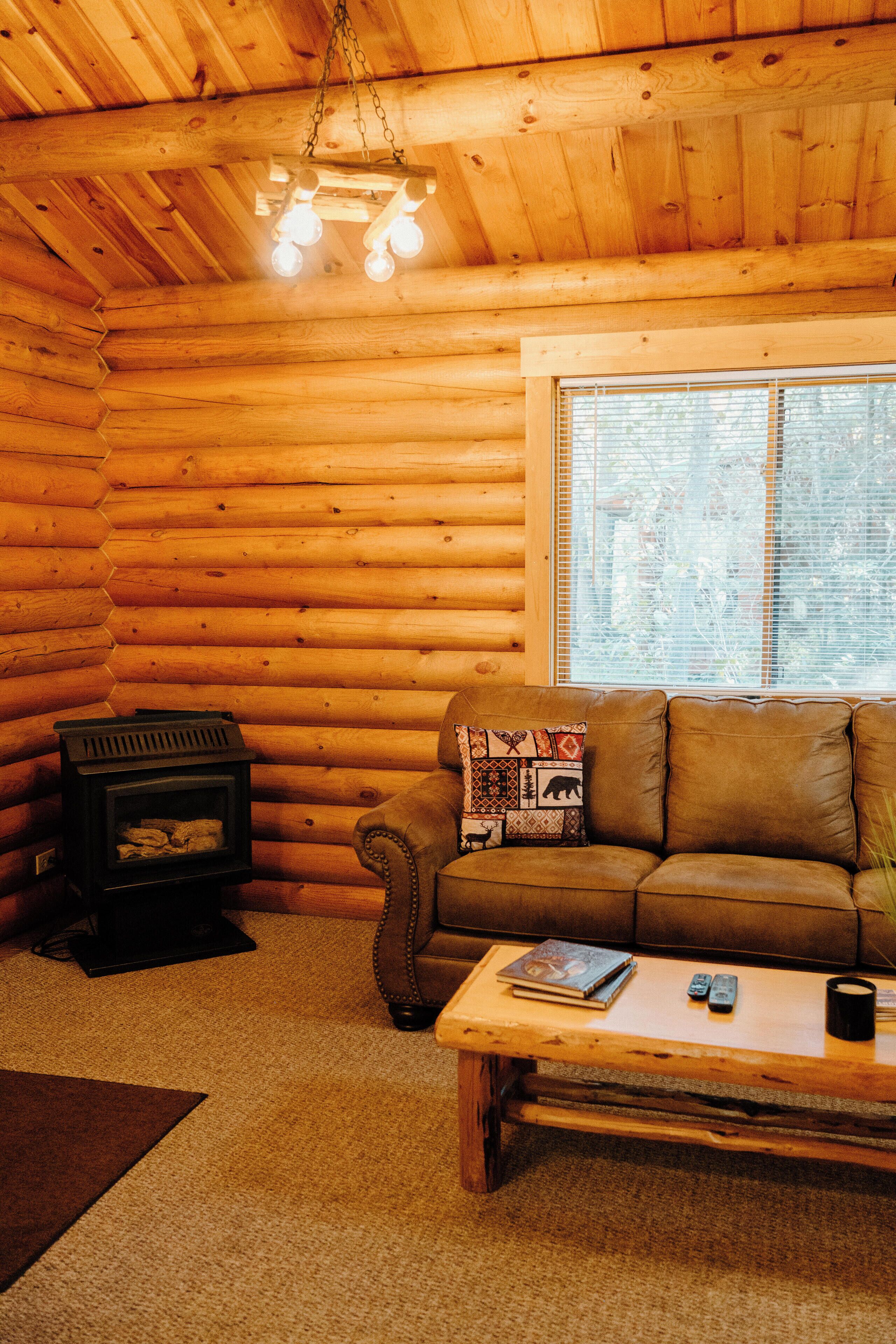 L’intérieur d’un chalet confortable, avec des murs en bois, un canapé en cuir, une table basse en bois, une cheminée et une fenêtre équipée de stores.