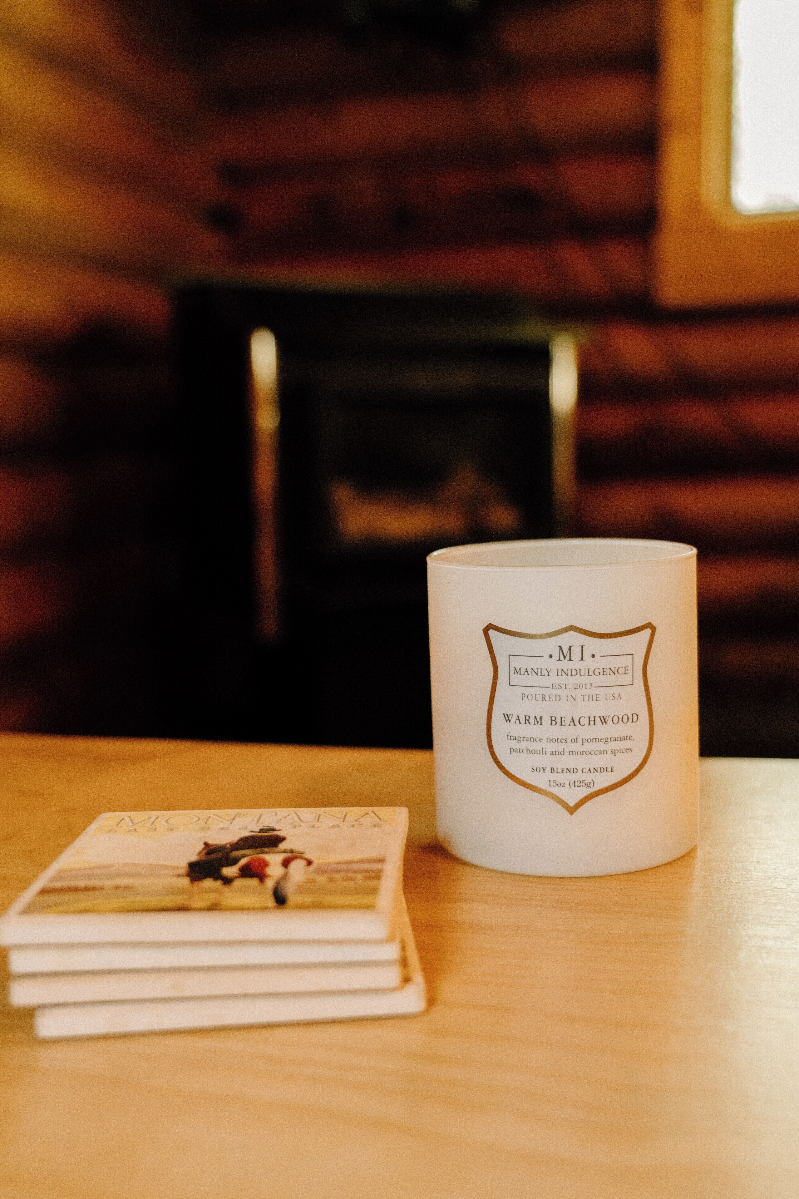 A white candle labeled "MI Manly Indulgence Warm Beachwood" and a stack of books on a wooden surface.
