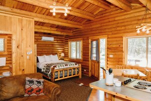 A log cabin interior with a wooden ceiling, a bed with a plaid bedspread, a wooden dining table, and a kitchen area with a stove.