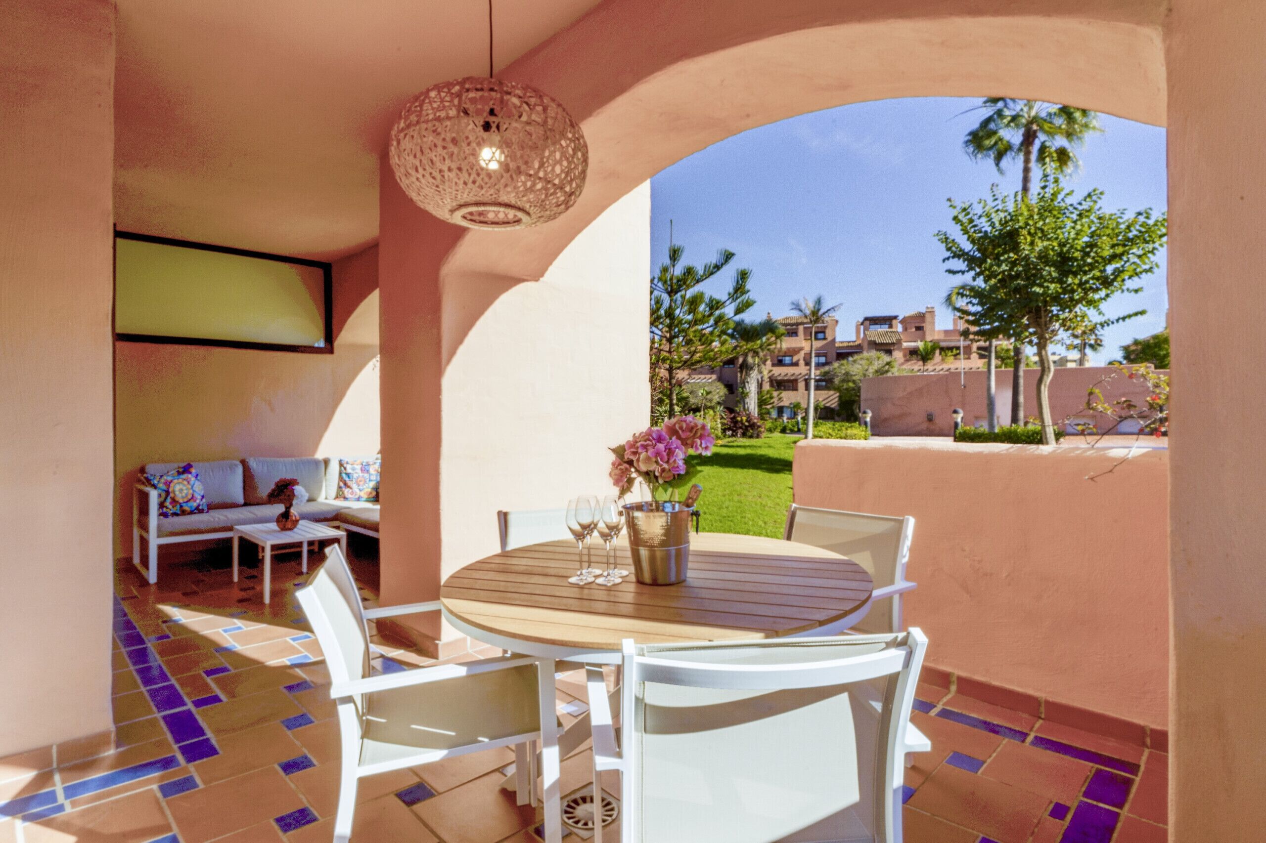 A patio with a round wooden table, white chairs, and a hanging light fixture.